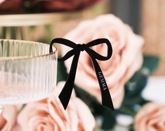 May include: A black acrylic bow-shaped glass charm featuring the name GEMMA engraved in white lettering on one of the ribbon tails. The charm is attached to the rim of a clear, vertically ribbed glass bowl with a gold-toned metallic edge.