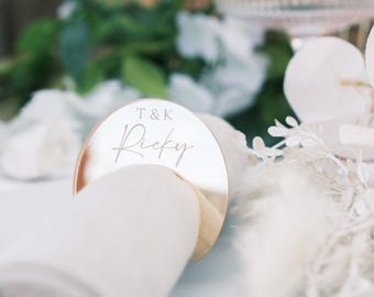 May include: A gold-colored, circular place card holder with the name "Ricky" engraved on it. The place card holder is positioned over a rolled-up white napkin. The text "T & K" is above the name. The background includes white flowers and greenery.