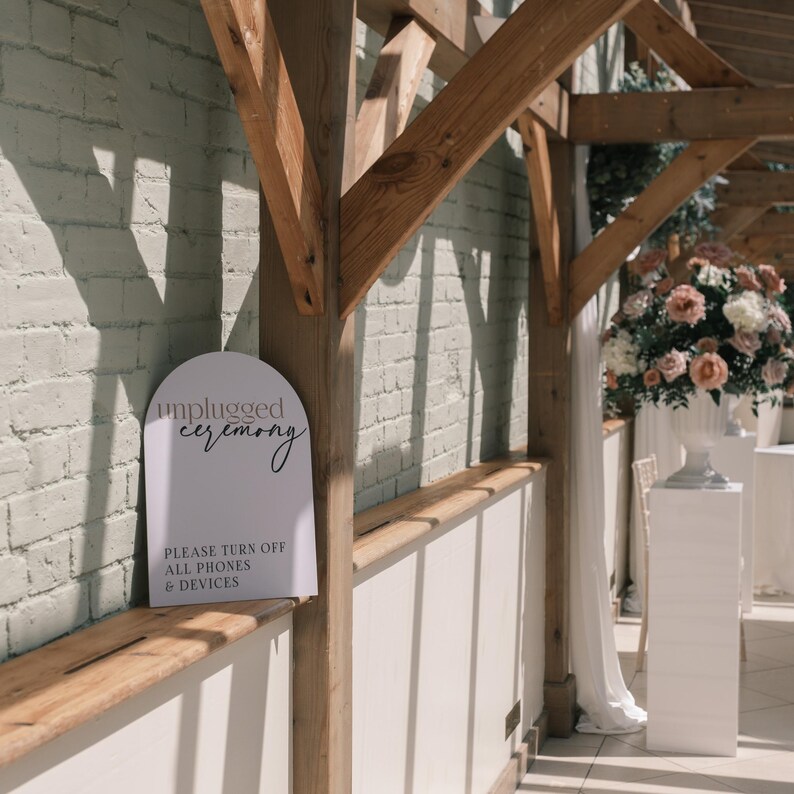 May include: A white sign with the words "unplugged ceremony" in elegant script. Below, it reads "Please turn off all phones & devices." The sign is set against a white brick wall and wooden beams, suggesting a wedding or event setting.