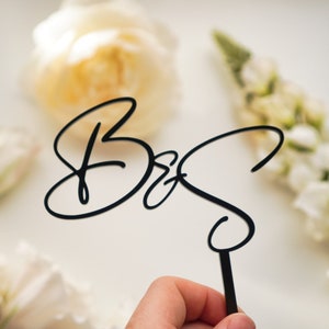 May include: Black acrylic cake topper with the letters "B & S" in a cursive script.