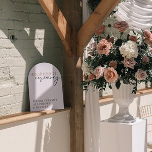May include: A white sign with the words "unplugged ceremony" and "Please turn off all phones & devices" in elegant script. The sign is placed near a wooden beam and a large floral arrangement with pink and white roses.