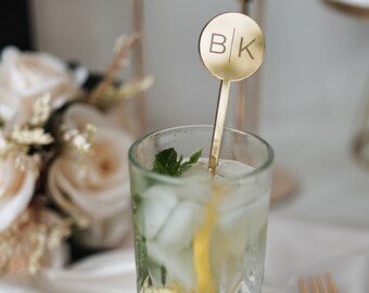 May include: A gold-toned metal cocktail stirrer featuring a circular top engraved with the letters B and K separated by a vertical line. The stirrer is placed inside a tall, clear glass filled with ice, a lemon slice, and fresh mint leaves.