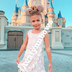 Puede incluir: Un vestido cruzado beige sin mangas con un patr&oacute;n repetitivo de cabezas de Mickey Mouse en azul claro y un dobladillo festoneado, combinado con orejas de rat&oacute;n con purpurina en oro rosa con un lazo central. El texto Avalynnbows es visible.