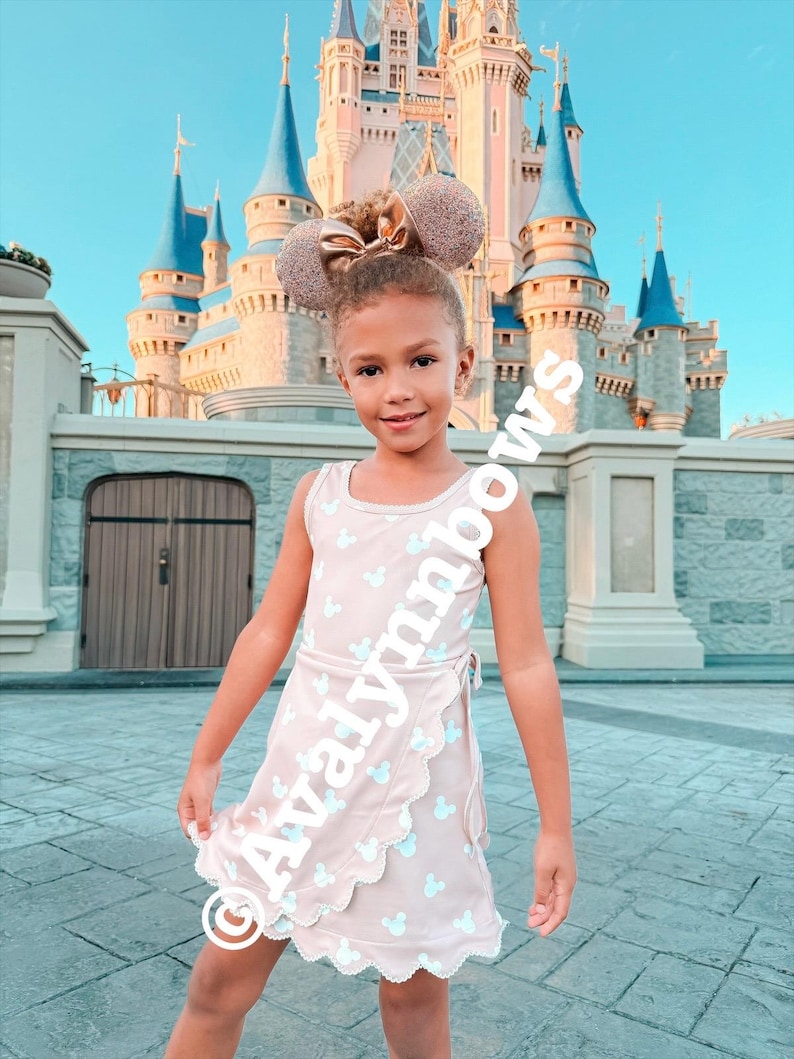Puede incluir: Un vestido cruzado beige sin mangas con un patr&oacute;n repetitivo de cabezas de Mickey Mouse en azul claro y un dobladillo festoneado, combinado con orejas de rat&oacute;n con purpurina en oro rosa con un lazo central. El texto Avalynnbows es visible.