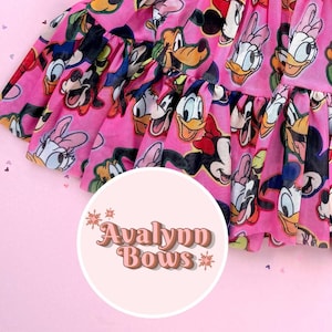 May include: A pink tulle skirt with a repeating pattern of cartoon characters such as Mickey and Minnie Mouse, Donald and Daisy Duck. The skirt has a ruffled hem and the text "Avalynn Bows" in a circular logo.