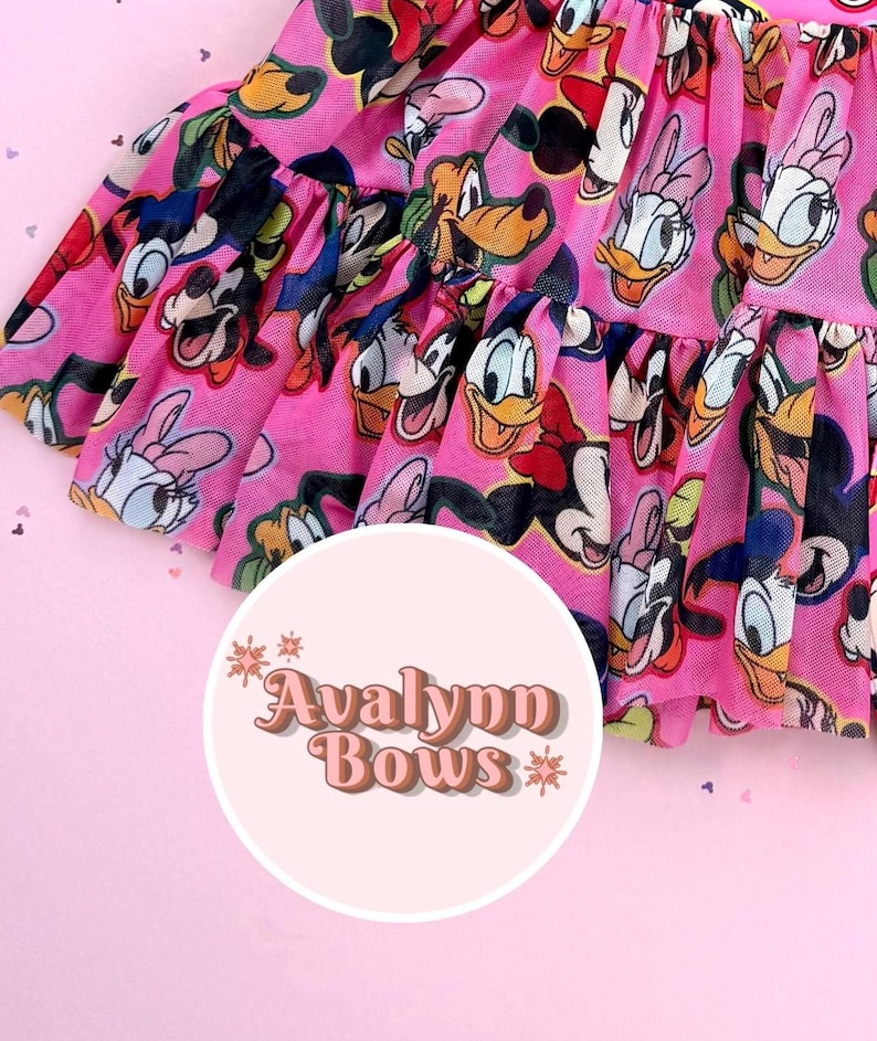 May include: A pink tulle skirt with a repeating pattern of cartoon characters such as Mickey and Minnie Mouse, Donald and Daisy Duck. The skirt has a ruffled hem and the text "Avalynn Bows" in a circular logo.