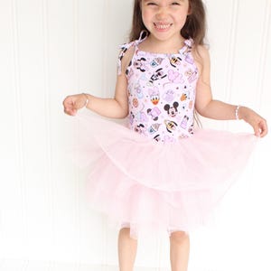 Può includere: Una bambina che indossa una gonna di tulle rosa e un vestito bianco senza maniche con una stampa di cartoni animati rosa e bianca. Il vestito ha sottili spalline che si legano alle spalle.