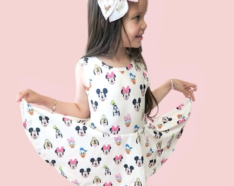 Könnte beinhalten: Ein junges Mädchen trägt ein weißes, ärmelloses Kleid mit einem Aufdruck verschiedener Disney-Figuren, darunter Mickey Mouse, Minnie Mouse, Donald Duck und Goofy. Sie trägt eine rosa-goldene Schleife im Haar.