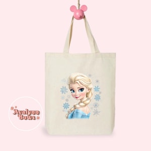 May include: A cream-coloured tote bag with a portrait of a blonde woman in a blue dress and snowflakes. The bag has long handles and is hanging from a pink Mickey Mouse-shaped hook. The bag has the text "Avalynn Bows" in a circle.
