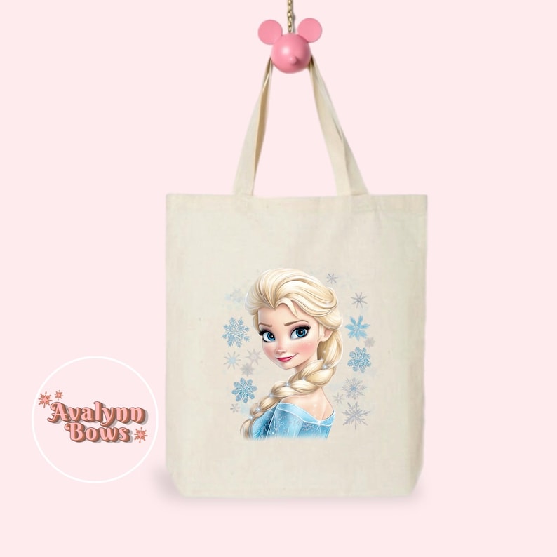 May include: A cream-coloured tote bag with a portrait of a blonde woman in a blue dress and snowflakes. The bag has long handles and is hanging from a pink Mickey Mouse-shaped hook. The bag has the text "Avalynn Bows" in a circle.