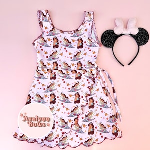 May include: A white sleeveless dress with a repeating pattern of Jasmine and Aladdin on a magic carpet, gold lamps, and small red flowers, featuring a scalloped hem and a matching headband with black glitter ears and a white bow. Text reads Avalynn Bows.