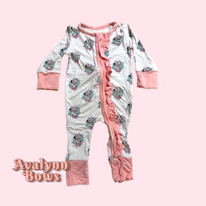 May include: White baby one-piece pajama with a repeating Minnie Mouse pattern. The pajama has long sleeves and a zipper down the front, accented with a pink ruffle. Pink trim is on the cuffs, ankles, and neckline. The text "Avalynn Bows" is visible.