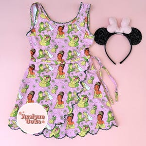 May include: A sleeveless light pink dress featuring a repeating pattern of a princess in a green gown, a frog, and white daisies, with dark green scalloped trim. A black headband with glittery ears and a light pink bow is included.