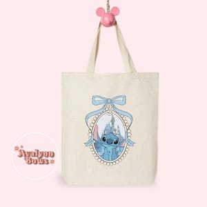 May include: A cream tote bag with a blue cartoon character and castle scene in a blue-ribboned oval frame. The bag hangs from a pink mouse-shaped hook. The text "Avalynn Bows" is on the lower left. The bag has two handles.
