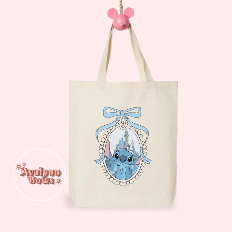 May include: A cream tote bag with a blue cartoon character and castle scene in a blue-ribboned oval frame. The bag hangs from a pink mouse-shaped hook. The text "Avalynn Bows" is on the lower left. The bag has two handles.