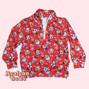 May include: Red pullover sweatshirt with a white zipper and a pattern of Disney characters and snowflakes. The sweatshirt features Mickey Mouse, Donald Duck, and other characters. The text "Avalynn Bows" is at the bottom.
