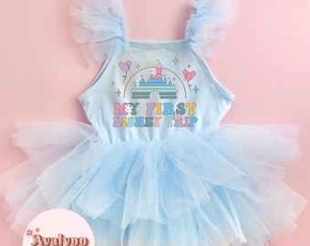 May include: Light blue tutu dress with the text "MY FIRST DISNEY TRIP" and a castle graphic. The dress features a layered tulle skirt and tulle shoulder straps. The background is a solid pink.