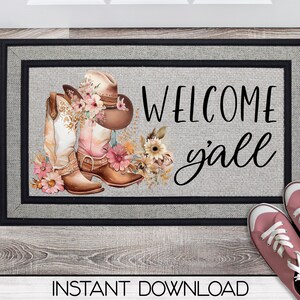 May include: A welcome mat with a gray background and black border. The text "Welcome y'all" is written in black cursive script. The mat is decorated with a floral design featuring pink, white, and brown cowboy boots and a brown cowboy hat.