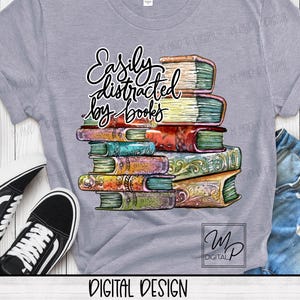 Puede incluir: Una camiseta gris con el texto "Easily distracted by books" en letra negra. Debajo del texto se muestra una pila de libros coloridos e ilustrados.