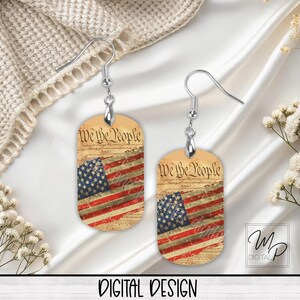 May include: A pair of silver earrings with a dog tag design. The dog tags feature a vintage-style American flag with the words "We the People" printed on the top.
