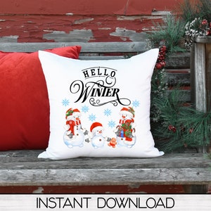 May include: White square pillow with a winter theme. The pillow features three snowmen wearing hats and scarves, with the words "Hello Winter" above them. Blue snowflakes are scattered around the snowmen. The pillow is on a wooden bench with a red pillow behind it.