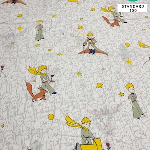 May include: White fabric with a repeating pattern featuring illustrations of a young prince, a fox, stars, and planets. The prince is depicted in various poses, wearing a yellow scarf and coat. The fabric has a "OEKO-TEX STANDARD 100" label.