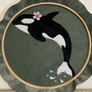 Könnte beinhalten: Gestickte schwarz-weiße Orca-Wal mit einer rosa Blume auf einem grünen runden Hintergrund.