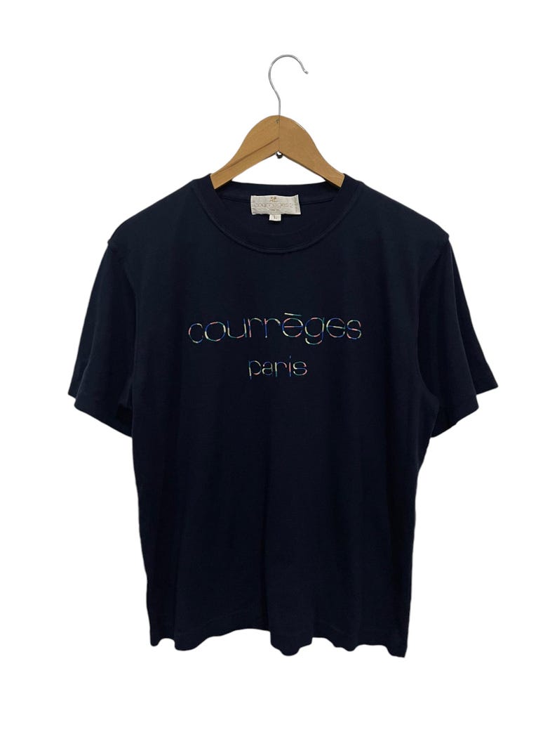 Op de afbeelding: Een marineblauw T-shirt met korte mouwen met een wit logo met de tekst "courr&egrave;ges paris" in een regenboogkleurig, iriserend lettertype.