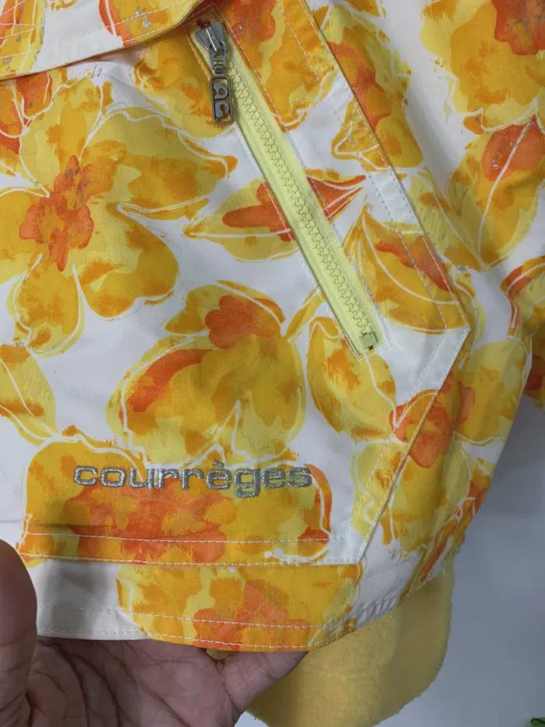 May include: White jacket with a vibrant floral pattern in yellow and orange. Features a yellow zipper and the brand name "courr&egrave;ges" in silver lettering. The jacket has a soft, yellow lining.