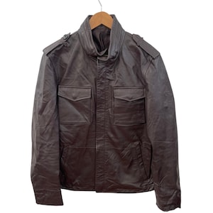 May include: A dark brown leather jacket with a high collar and two front pockets. The jacket is suspended from a wooden hanger. The jacket features shoulder epaulettes and a zip closure.
