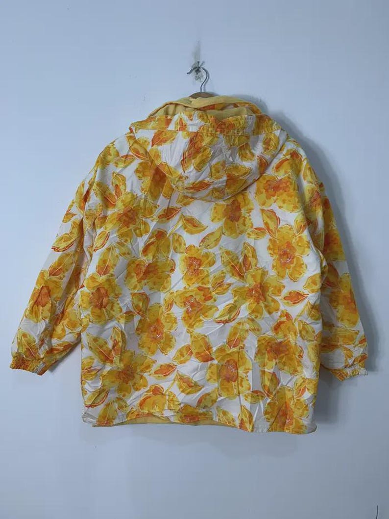 May include: A hooded jacket with a floral pattern in shades of yellow and orange on a white background. The jacket has long sleeves and a drawstring hood. The jacket is hanging on a metal hanger.