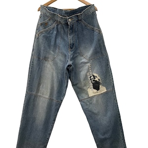 May include: Light blue denim jeans with a faded wash, featuring a patch design on the right leg with a black and white image and the text "MAKAVELI BRANDED". The jeans are hanging on a wooden hanger.