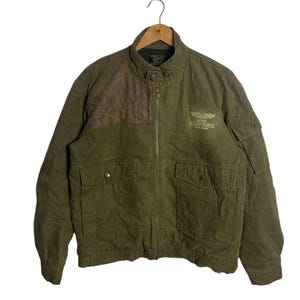 Peut inclure: Veste vert olive avec un patch en cuir marron sur l'épaule. La veste a une fermeture éclair, plusieurs poches et le texte "US AIR FORCE" avec un emblème d'aile. La veste est suspendue à un cintre en bois.