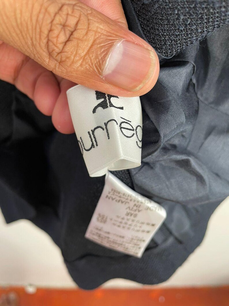May include: Close-up of a white label with the brand name "courr&egrave;ges" printed in black. The label is attached to a dark navy blue garment. The garment's lining is visible.