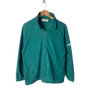 Puede incluir: Una chaqueta de cremallera de forro polar verde azulado con una etiqueta blanca en la manga que dice "Salomon Clothing" y un logotipo bordado verde en la parte delantera que dice "Salomon".