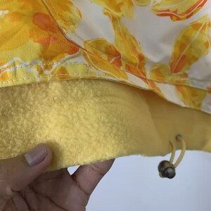 May include: Close-up of a jacket with a floral pattern in yellow and orange on a white background. The interior is a solid yellow fleece. A drawstring with wooden beads is visible.