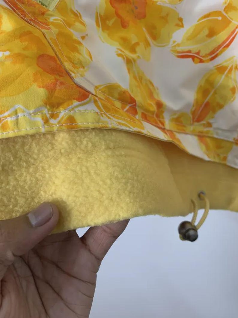 May include: Close-up of a jacket with a floral pattern in yellow and orange on a white background. The interior is a solid yellow fleece. A drawstring with wooden beads is visible.