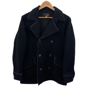 Peut inclure: Un caban noir à double boutonnage avec des détails de couture blancs. Le manteau a un col cranté, deux poches avant et des boutons noirs. Il est suspendu à un cintre en bois sur fond blanc.
