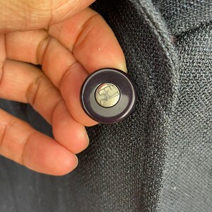 May include: Close-up of a dark gray button with a silver logo on a dark gray blazer. The button is round with a dark purple rim. The blazer has a textured fabric.
