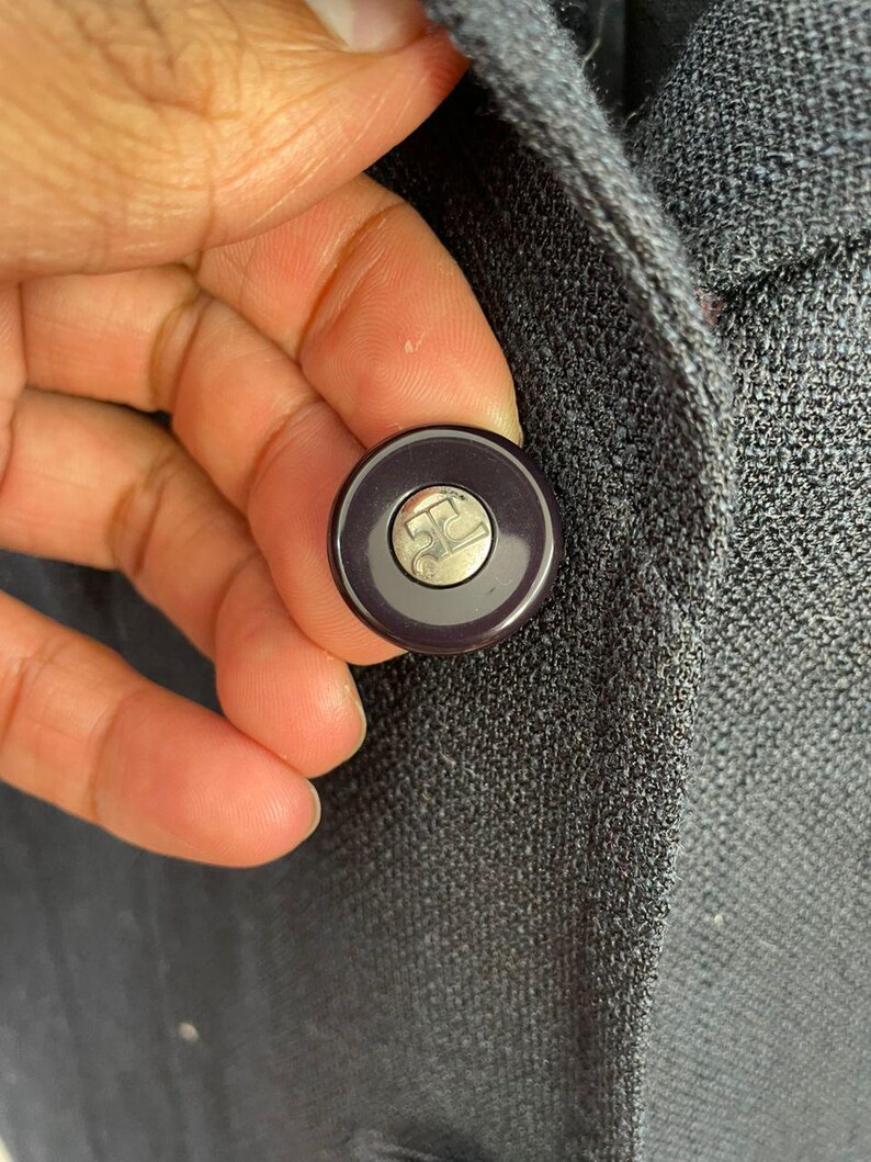 May include: Close-up of a dark gray button with a silver logo on a dark gray blazer. The button is round with a dark purple rim. The blazer has a textured fabric.