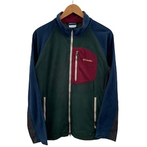 Könnte beinhalten: Mehrfarbige Fleecejacke mit durchgehendem Reißverschluss. Die Jacke hat einen dunkelgrünen Körper, marineblaue Ärmel und eine weinrote Brusttasche mit dem "Columbia"-Logo. Die Jacke hängt an einem Holzbügel.