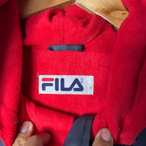 Pu&ograve; includere: Una giacca blu navy foderata in pile rosso con un'etichetta logo Fila. L'etichetta &egrave; bianca con lettere rosse e nere. L'etichetta dice "FILA" con un simbolo di marchio registrato e "J M" sotto.