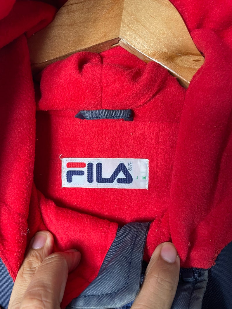 Pu&ograve; includere: Una giacca blu navy foderata in pile rosso con un'etichetta logo Fila. L'etichetta &egrave; bianca con lettere rosse e nere. L'etichetta dice "FILA" con un simbolo di marchio registrato e "J M" sotto.
