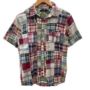 May include: Short-sleeved patchwork shirt in a variety of colors, including red, green, blue, and white. The shirt is made of a lightweight fabric and features a button-down front and a chest pocket. The shirt is hanging on a wooden hanger.