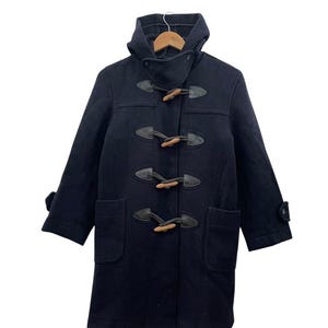 Pode incluir: Um casaco duffle azul marinho com capuz e fechos de alternância. O casaco apresenta dois bolsos frontais e punhos abotoados. As alternâncias são feitas de madeira e couro.
