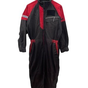 Op de afbeelding: Zwart en rood racepak met het Suzuki-logo op de mouw. Het racepak heeft een hoge kraag, een ritssluiting aan de voorkant en meerdere zakken. De schouders en de bovenborst zijn rood, de rest van het pak is zwart.