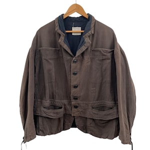 Könnte beinhalten: Braune Jacke mit dunkelblauem Kragen und schwarzen Knöpfen. Die Jacke hat zwei Vordertaschen und hängt an einem Holzbügel. Die Jacke scheint aus einem strapazierfähigen Stoff zu sein.