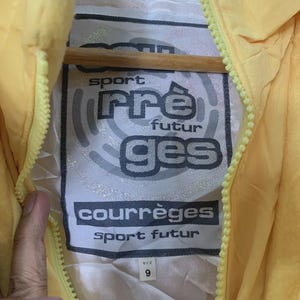 May include: Close-up of a yellow jacket with a zipper. The jacket's interior label features the text "sport pre futur ges courr&egrave;ges sport futur" in a black and white design. The size tag reads "9".