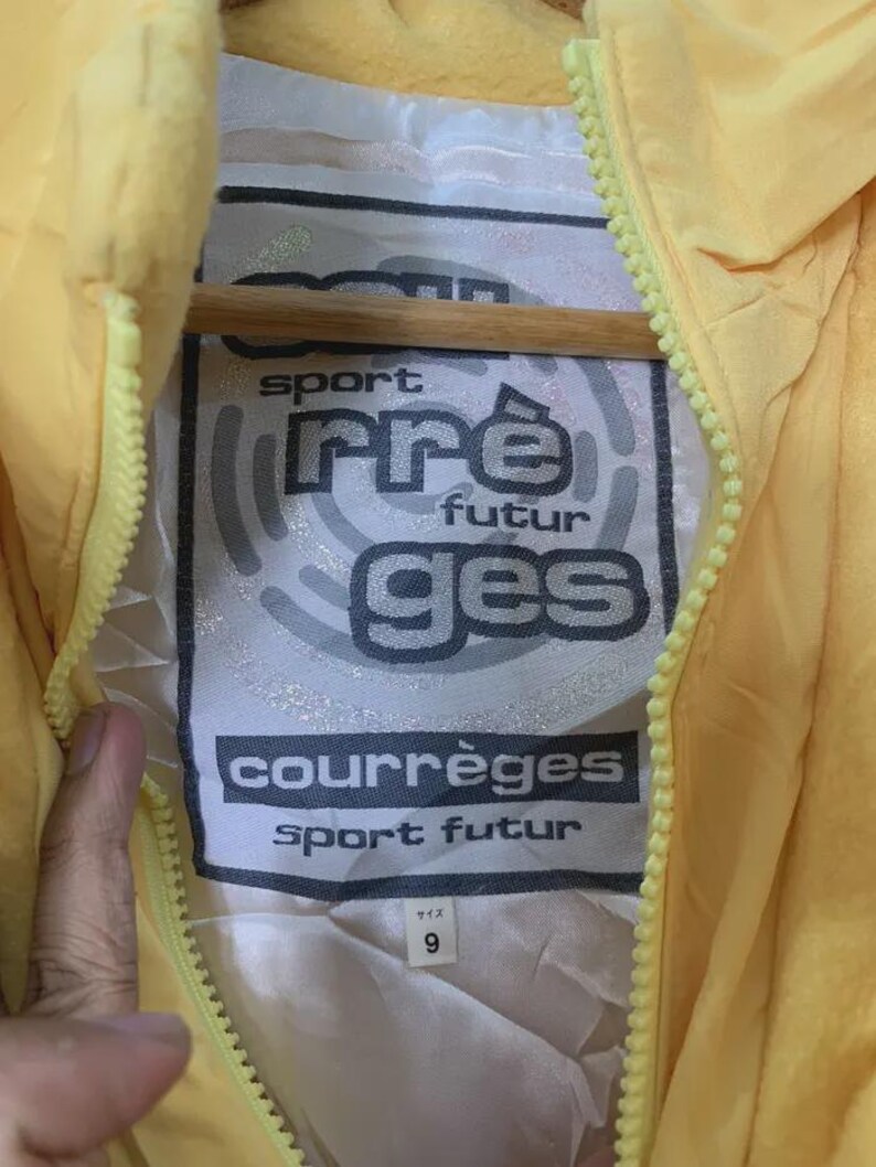 May include: Close-up of a yellow jacket with a zipper. The jacket's interior label features the text "sport pre futur ges courr&egrave;ges sport futur" in a black and white design. The size tag reads "9".