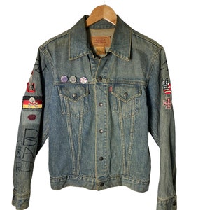 May include: A blue denim jacket with patches and pins on the sleeves. The jacket has a button-up front and a pointed collar. The patches include a skull and crossbones, a peace sign, and a red, white, and blue flag.